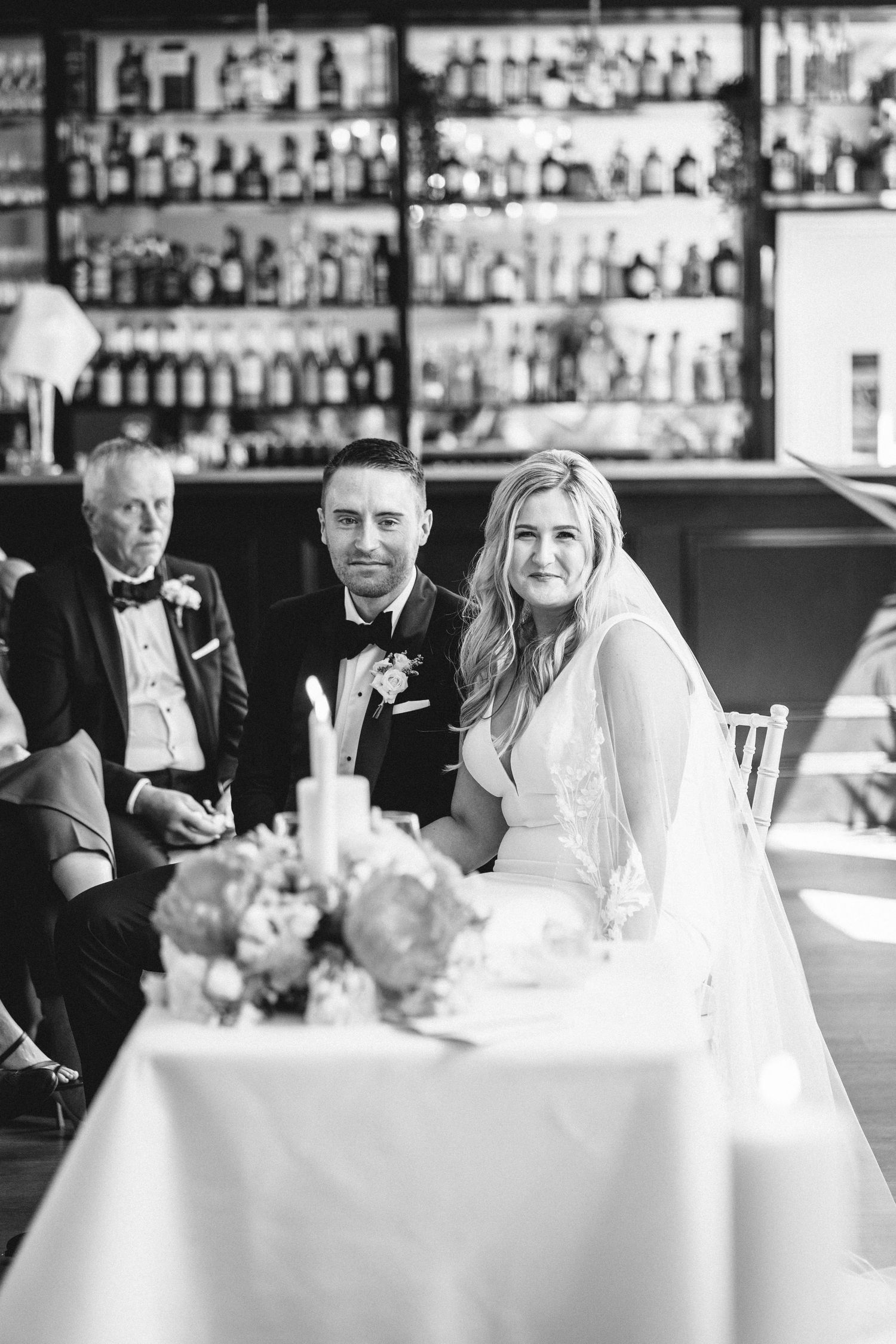 Wedding couple sitting at a reception table with a bar display of bottles in the background.
