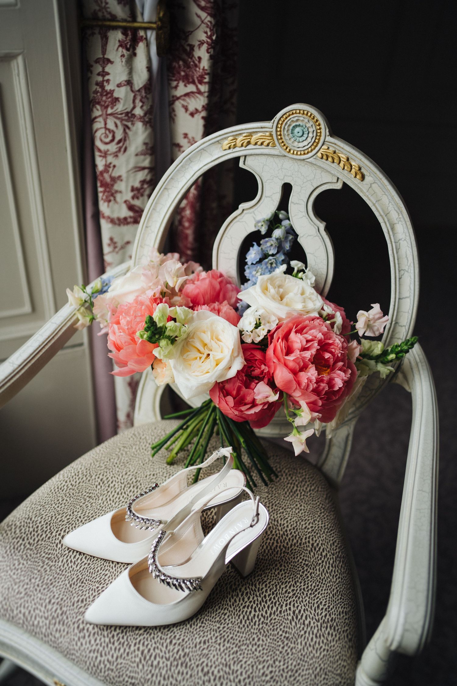 White bridal shoes and pink floral bouquet arranged on an ornate vintage chair with decorative mirror.