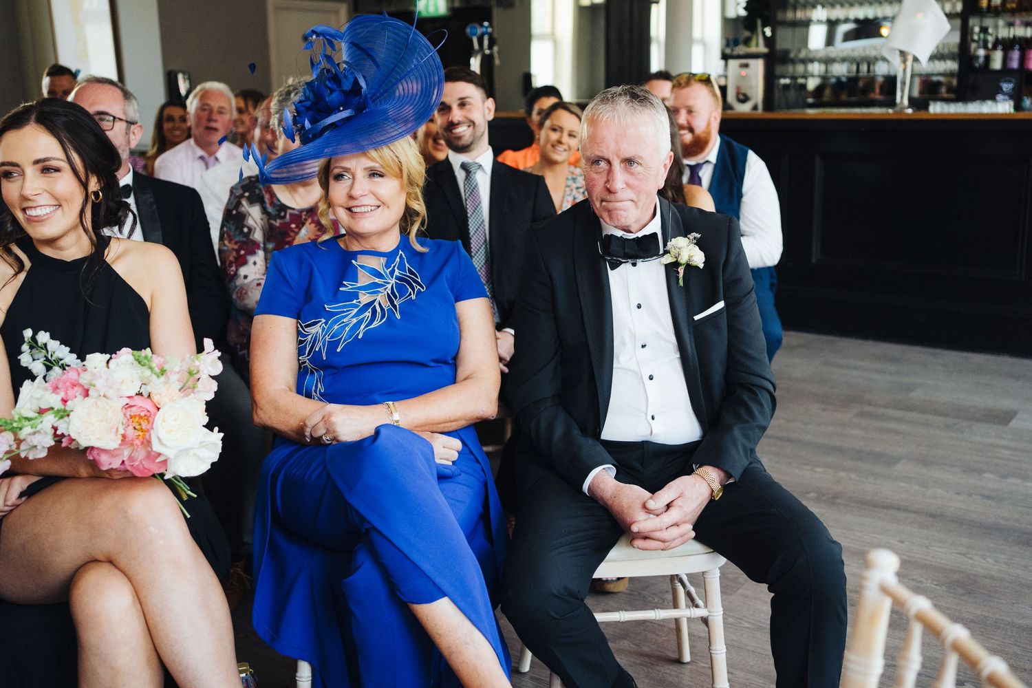 Wedding guests in formal attire including a striking blue outfit watch the ceremony from their seats.