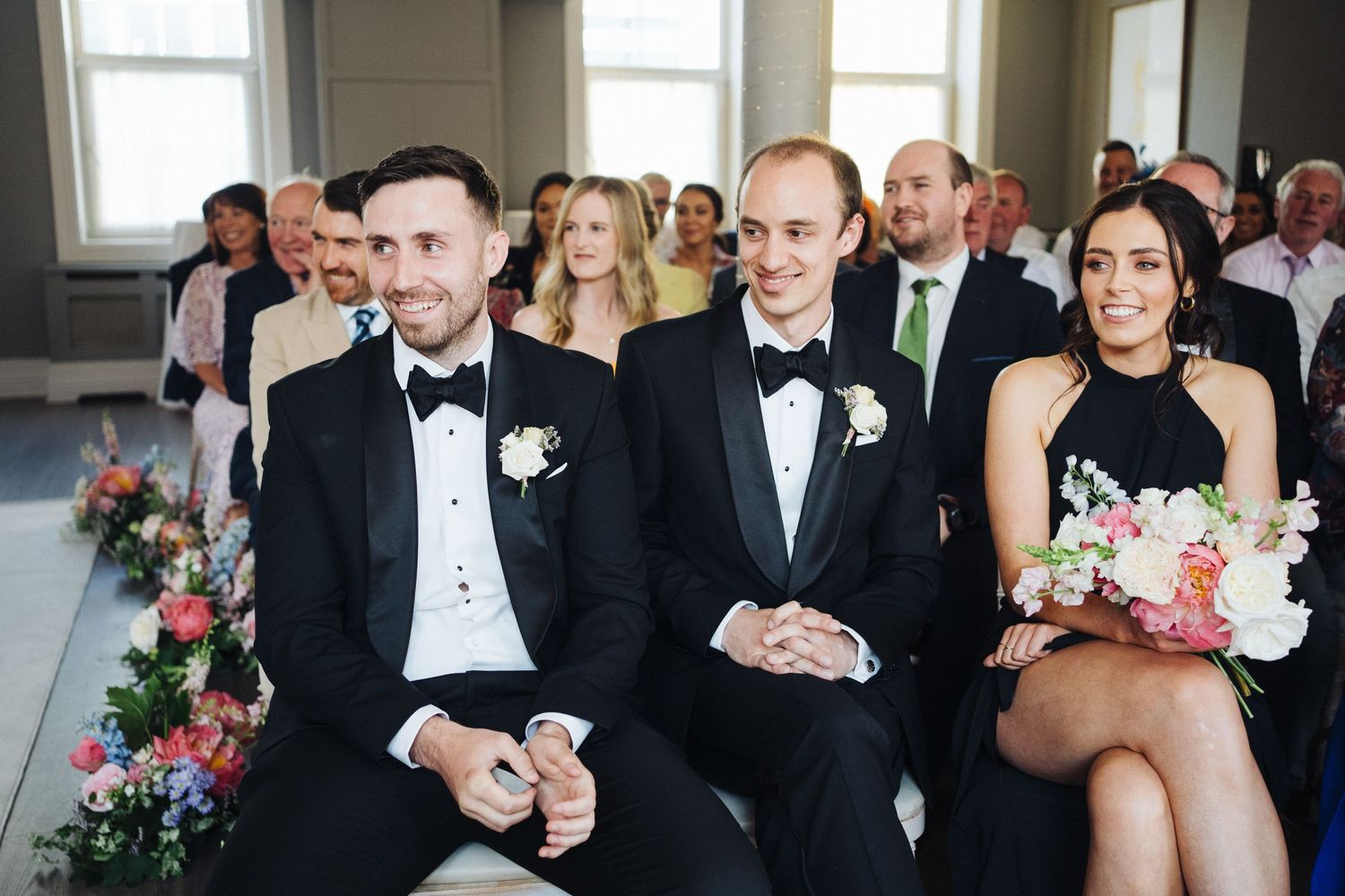 Wedding guests in formal black tie attire seated during the ceremony with floral arrangements visible.