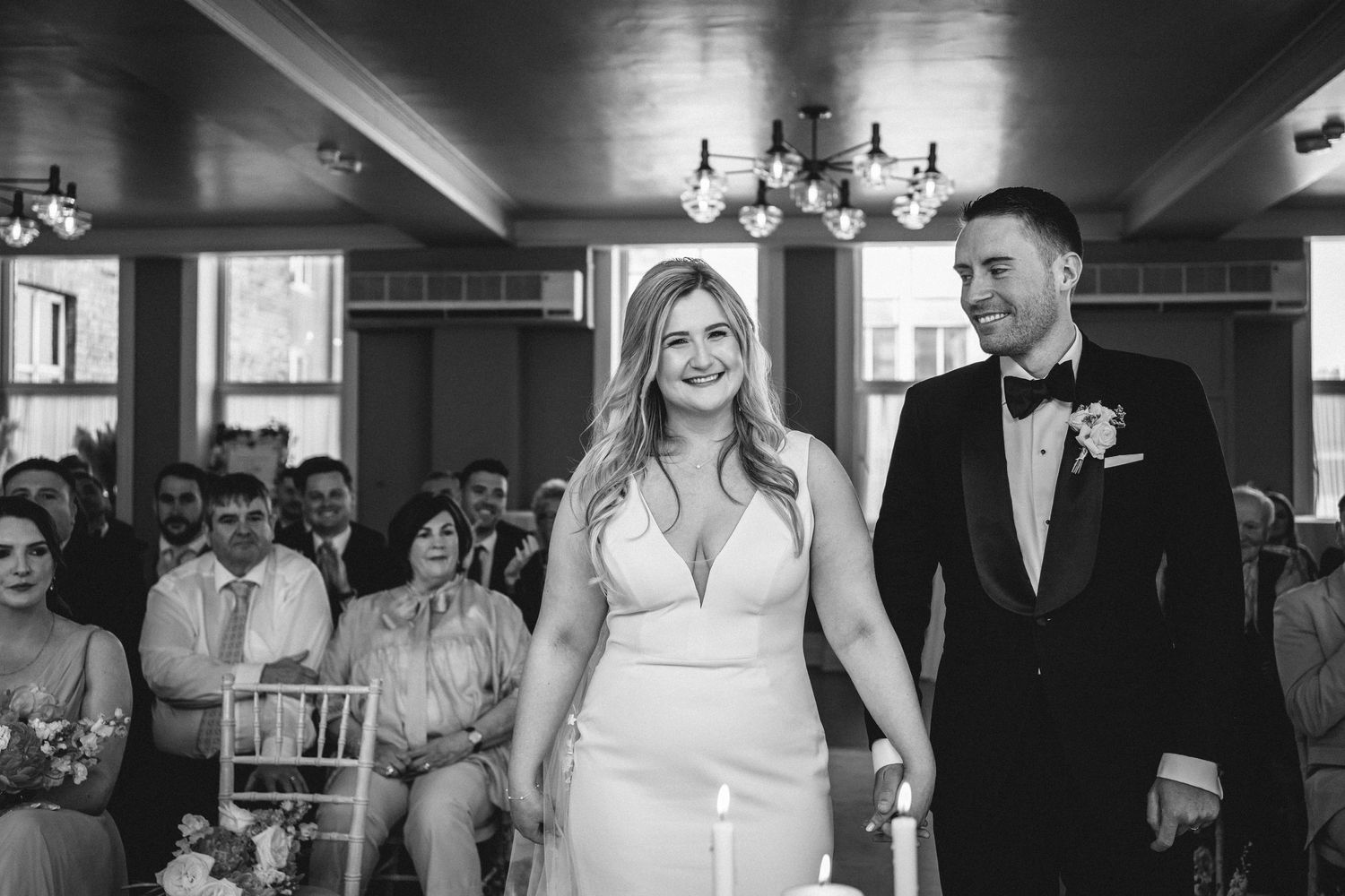 Newlyweds walk down the aisle together in black and white as guests look on in an elegant venue.