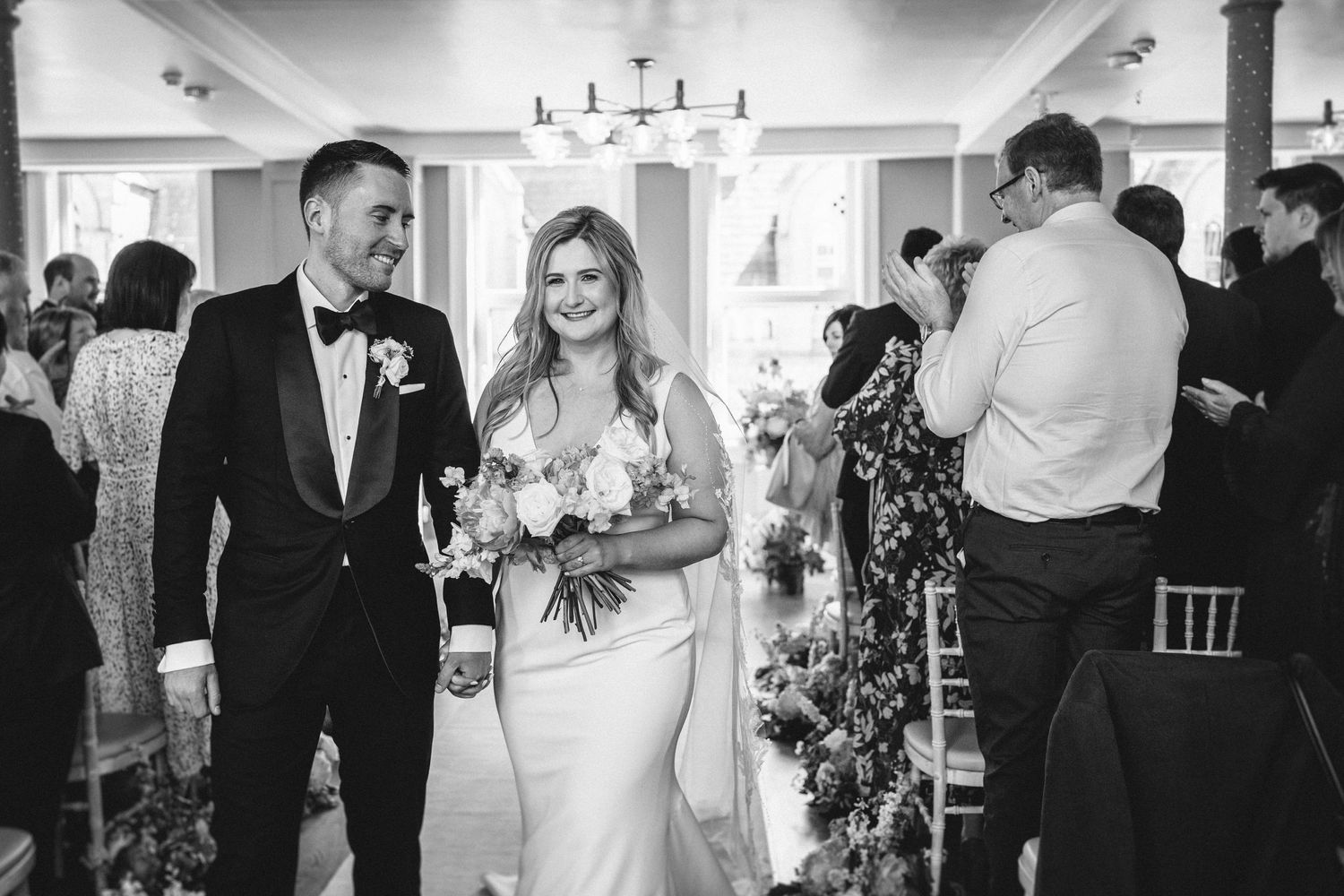 A black and white photo of newlyweds walking down the aisle while guests celebrate in background.