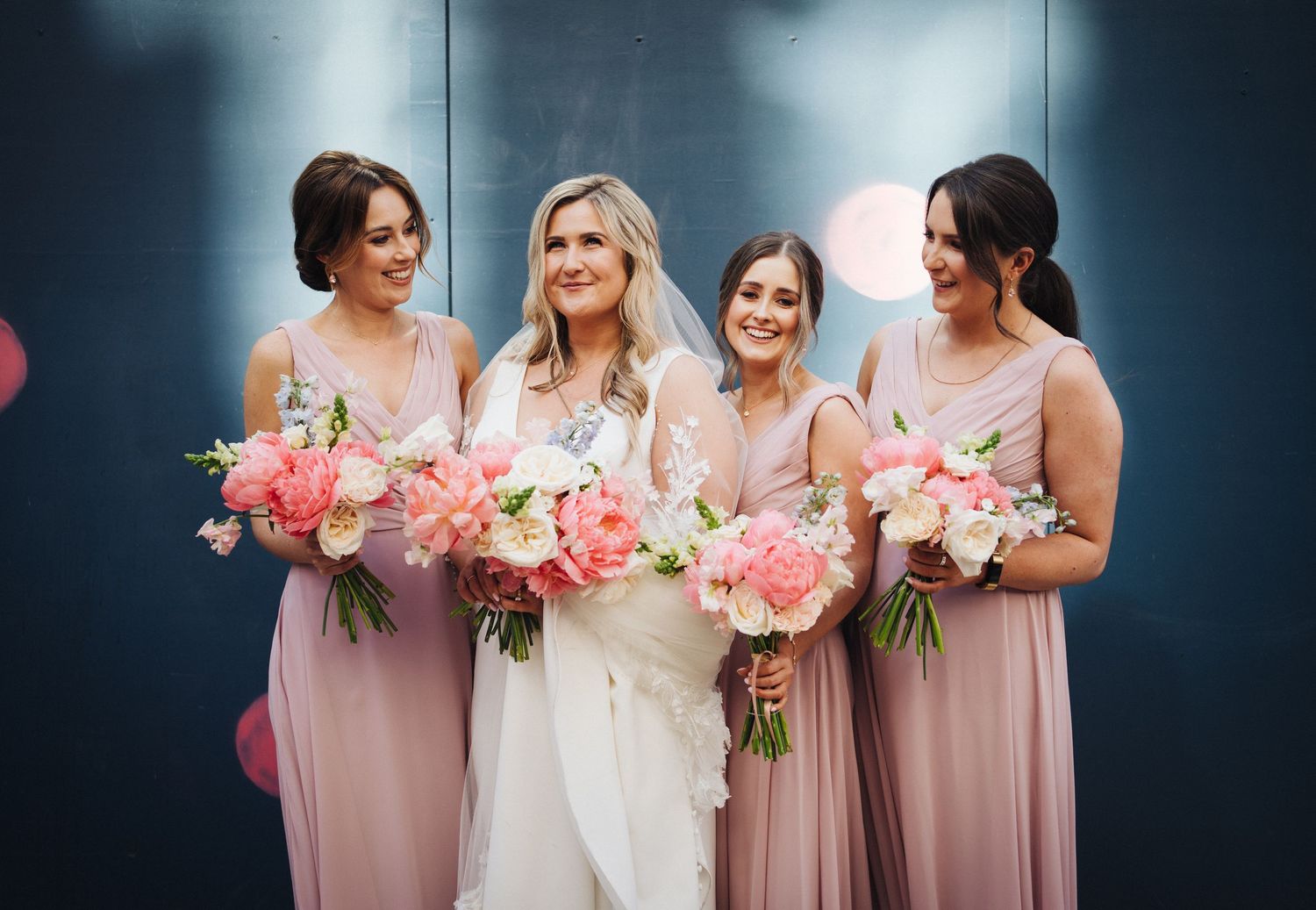 Wedding party in blush pink dresses hold pink and white bouquets against dark backdrop.