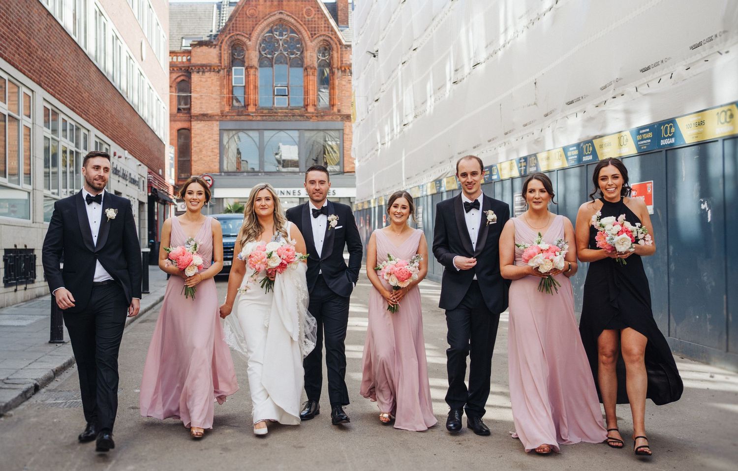 Wedding party poses outdoors in formal attire with pink bridesmaid dresses against urban backdrop.