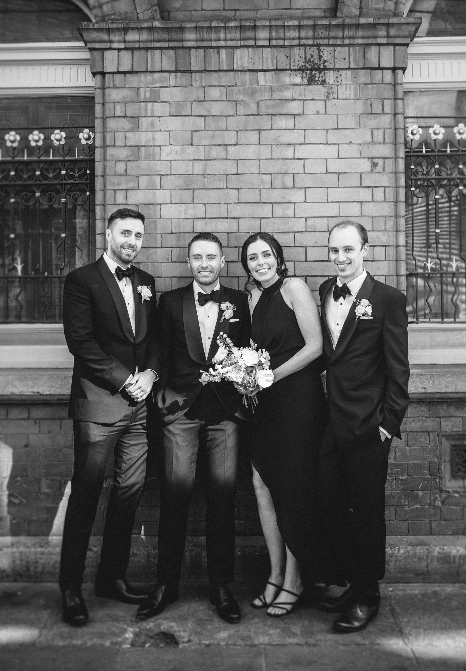 Black and white portrait of wedding party members in formal attire against brick building.