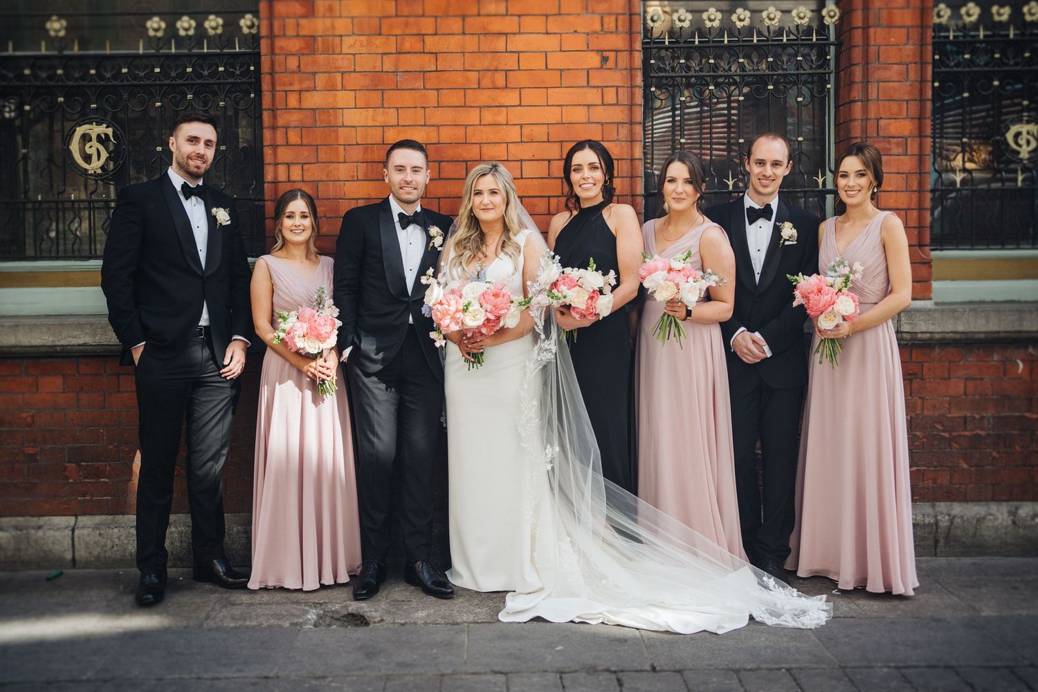 Wedding party in pink dresses and black tuxedos pose together against brick building.