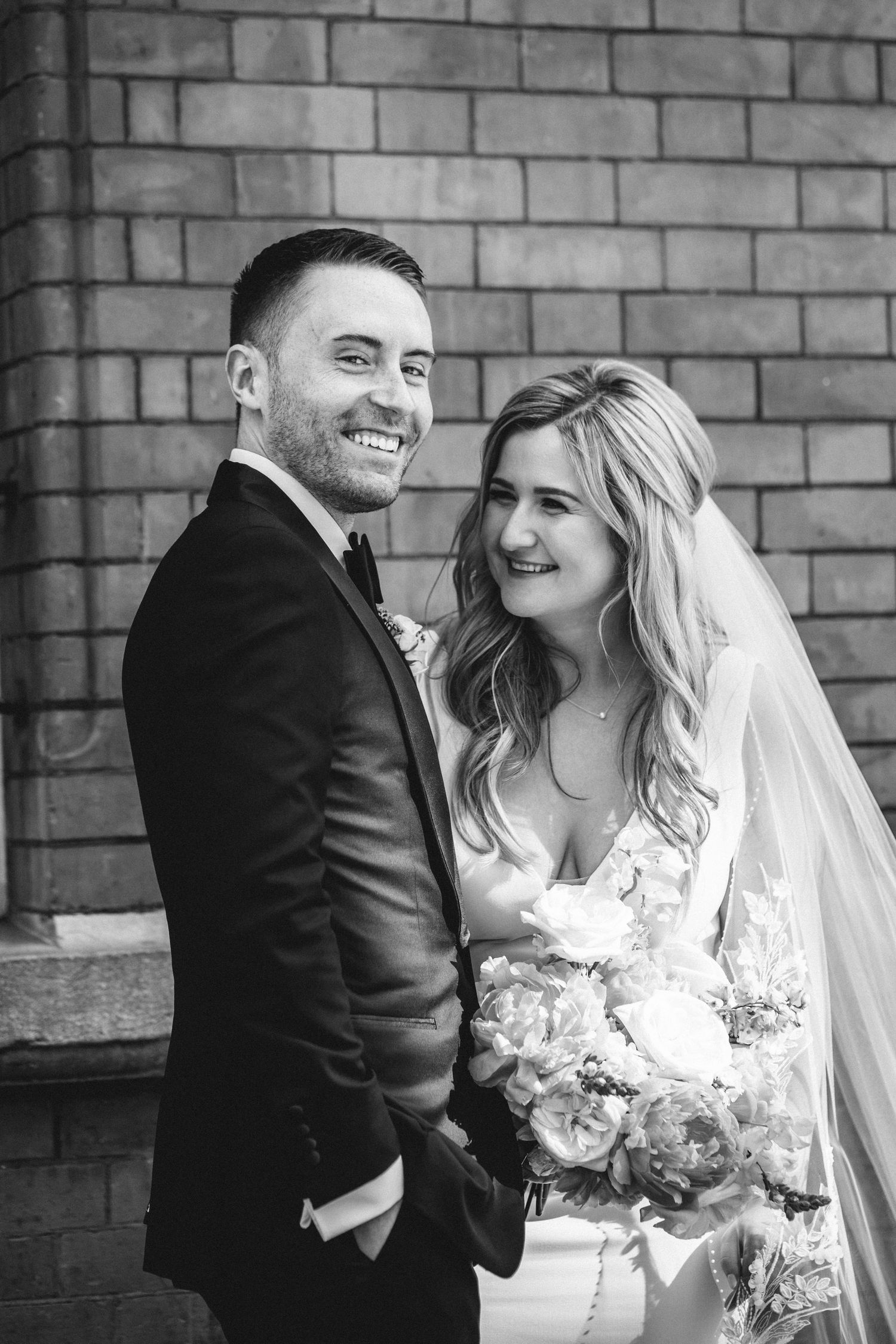 Black and white portrait of newlyweds smiling together against brick wall.