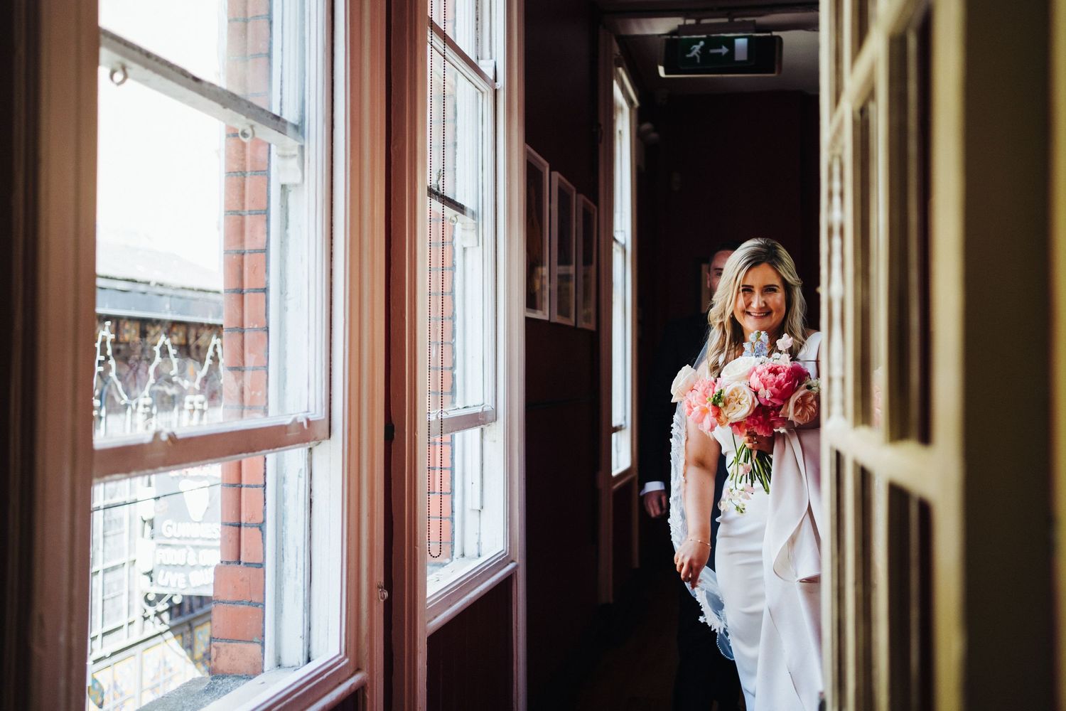 Bride holding bouquet poses by window in vintage hallway setting.