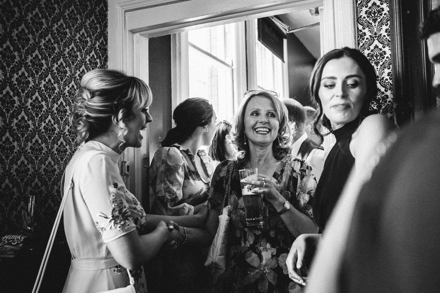 Black and white candid shot of wedding guests mingling and laughing in an elegant venue.