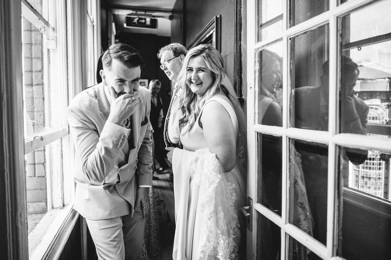 A black and white photo captures laughter and joy between wedding guests by window light.
