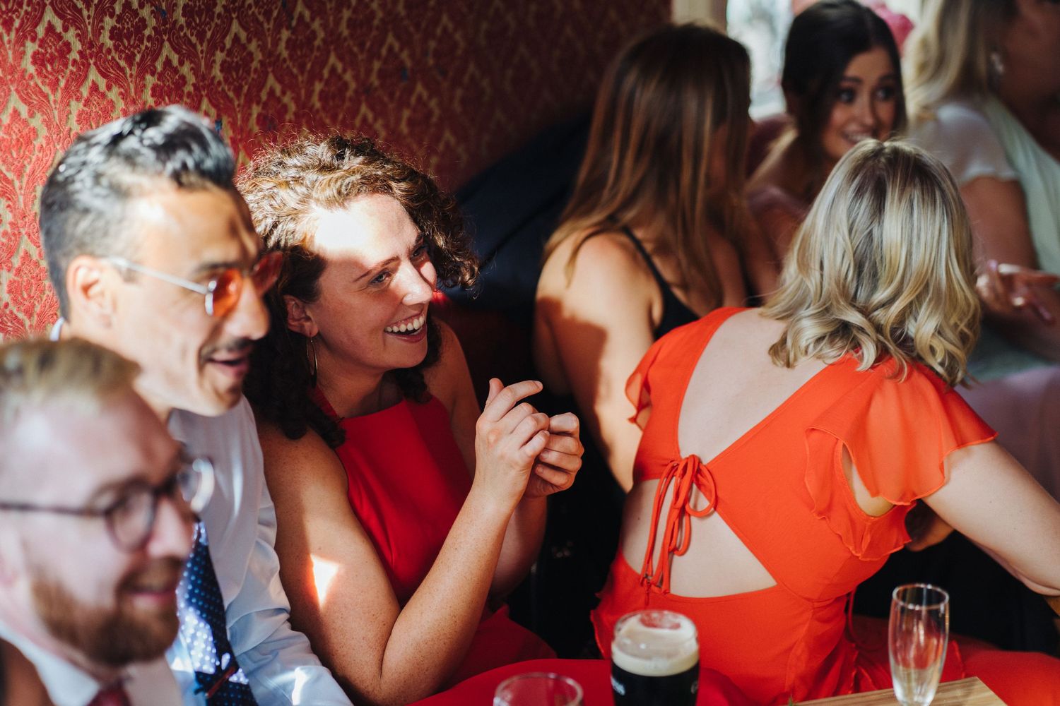 Wedding guests in bright attire share animated conversation over drinks at a red-walled venue.