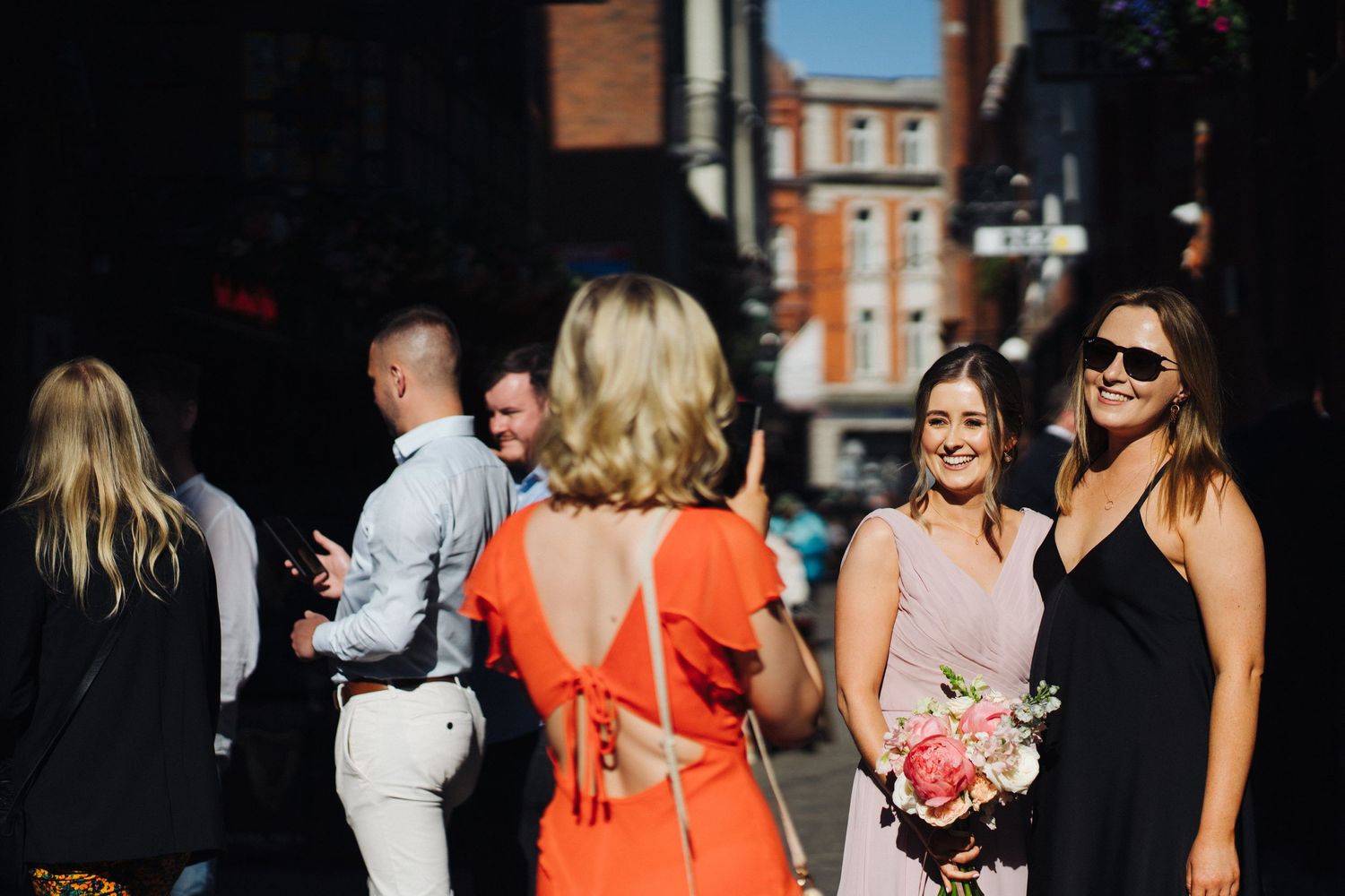 Wedding guests gather outdoors on a sunny day with one wearing a vibrant orange dress holding flowers.