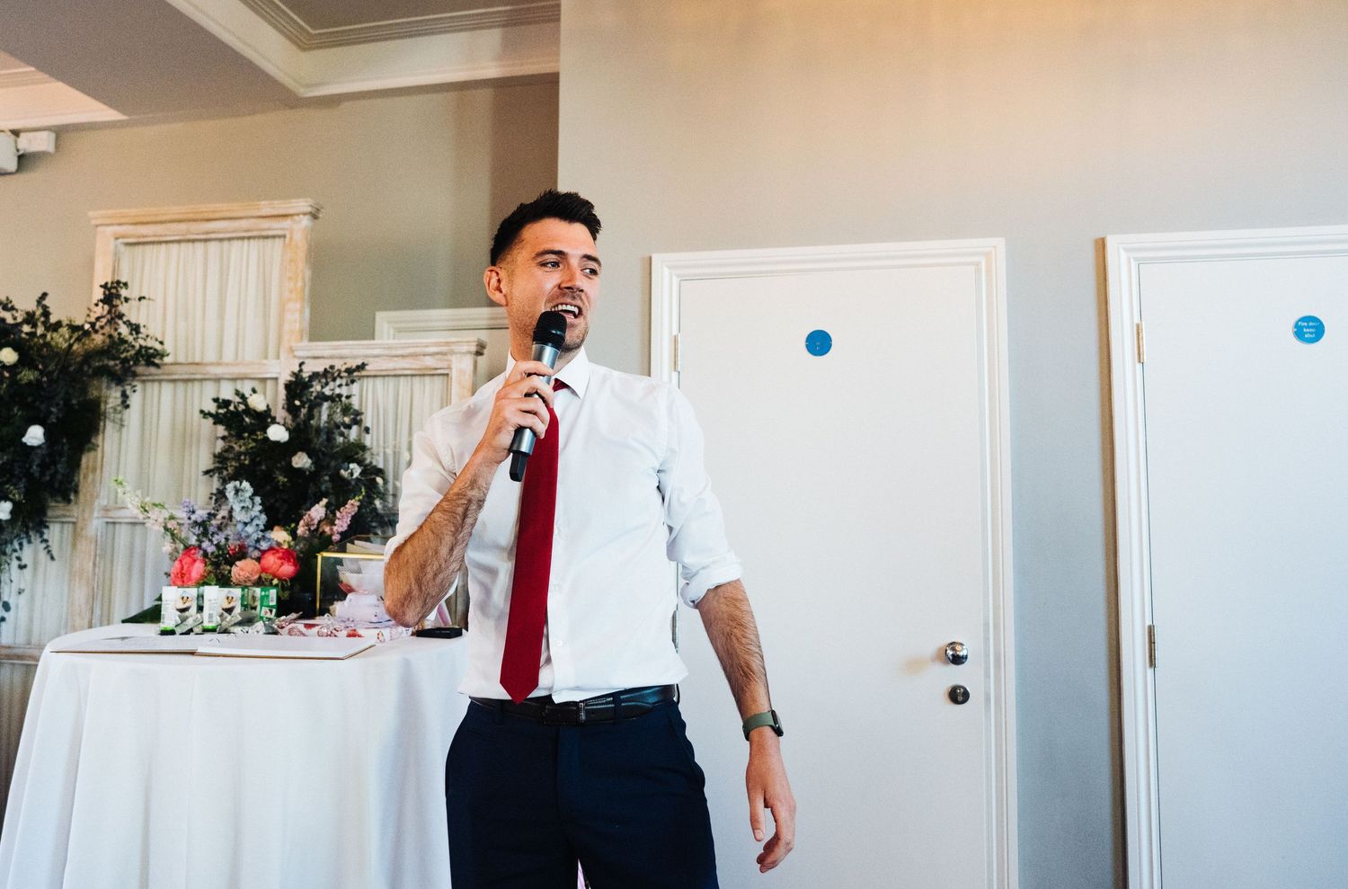 Speaker in white shirt and red tie delivers speech at wedding reception.