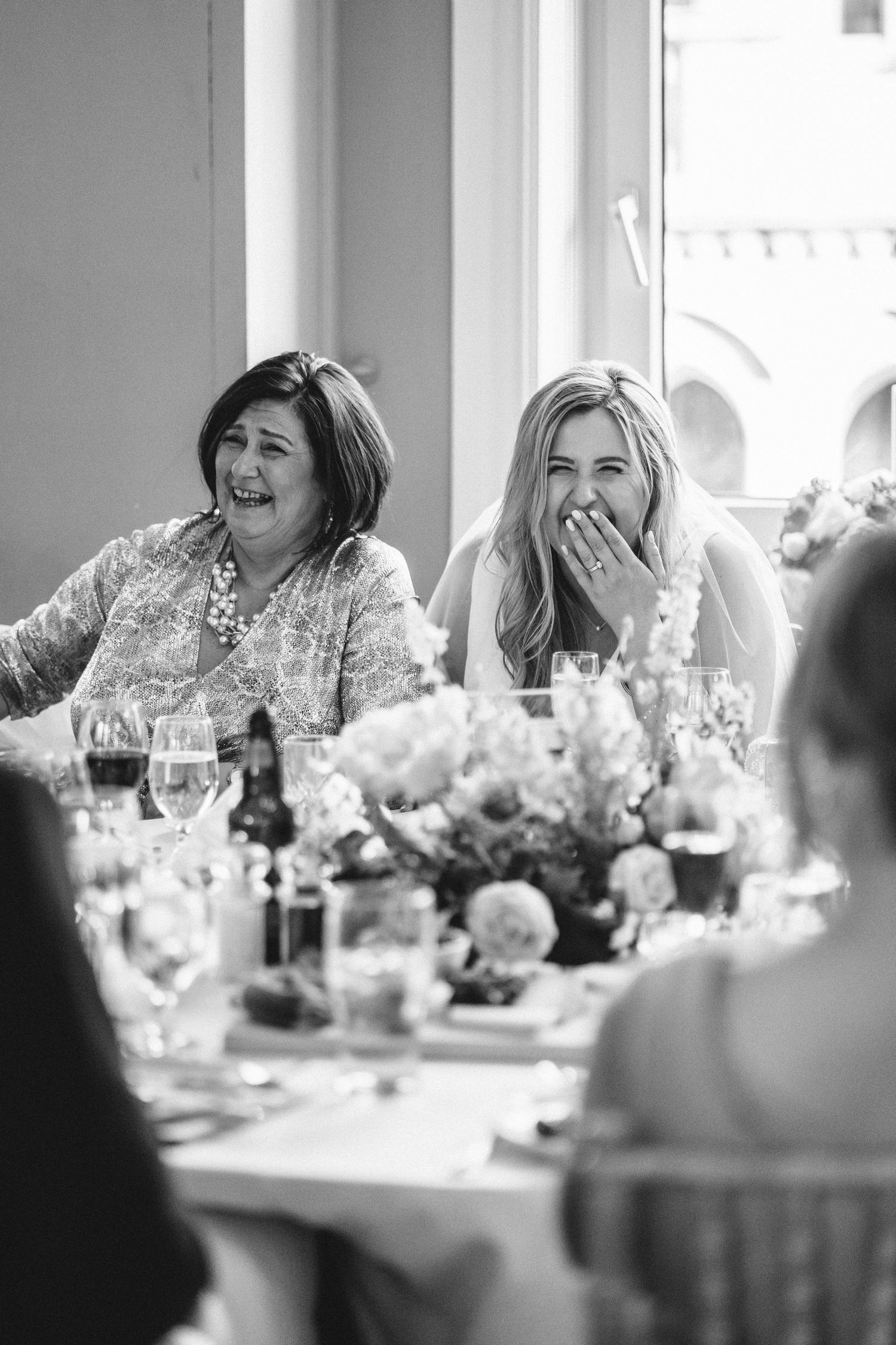 Wedding guests laugh joyfully at a decorated reception table with floral centerpieces.