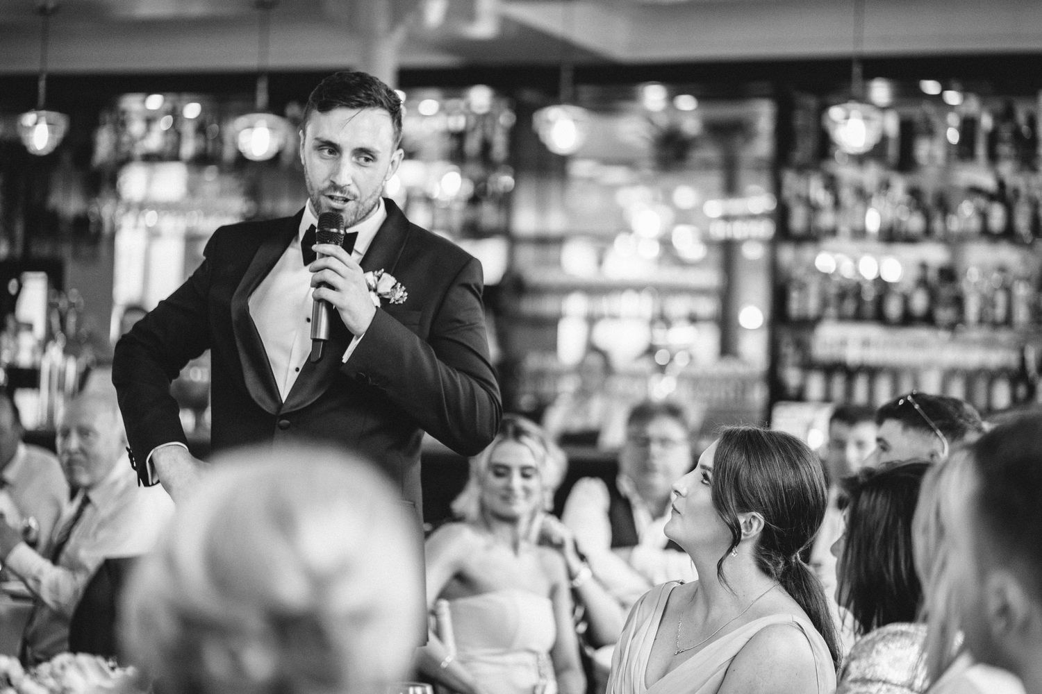 A speaker addresses wedding guests during a reception in a dimly lit venue with a bar backdrop.