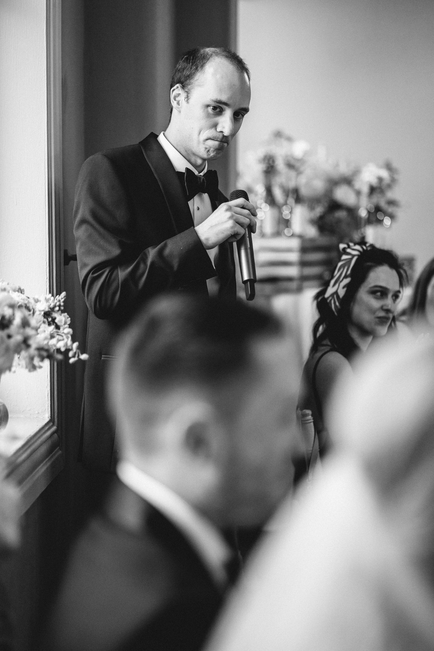 A speaker in a tuxedo delivers remarks near a window during a formal wedding celebration.