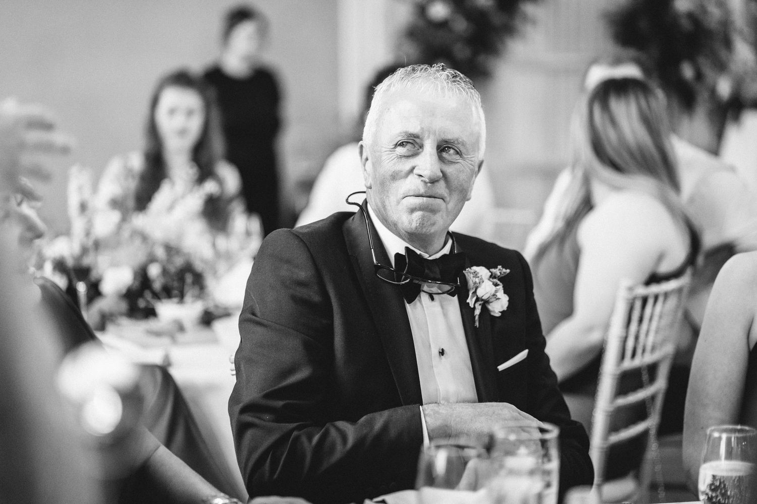 A guest in formal attire sits at an elegant wedding reception table wearing a black tuxedo and bowtie.