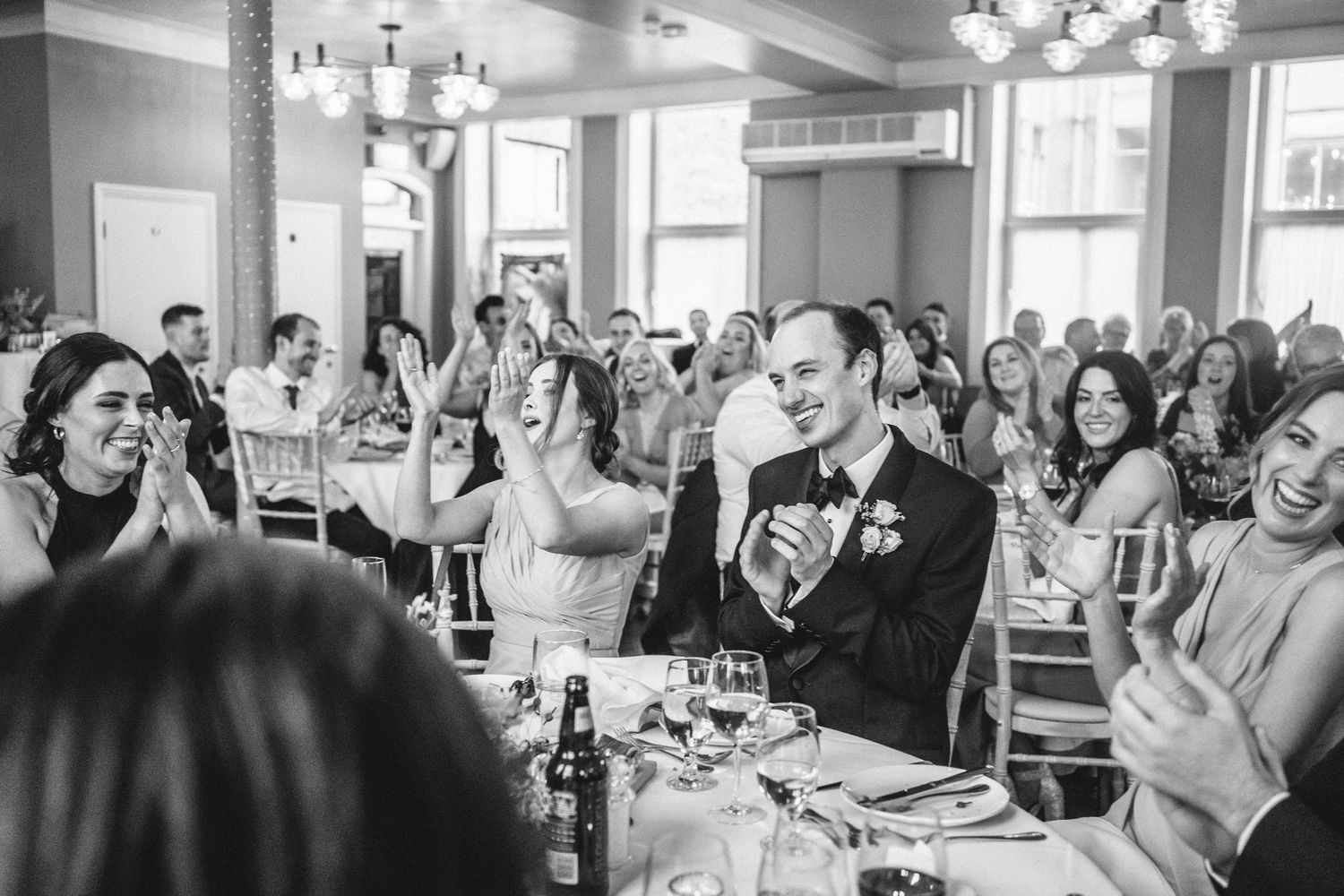 Wedding guests enthusiastically applaud during a reception speech in an elegant dining room.
