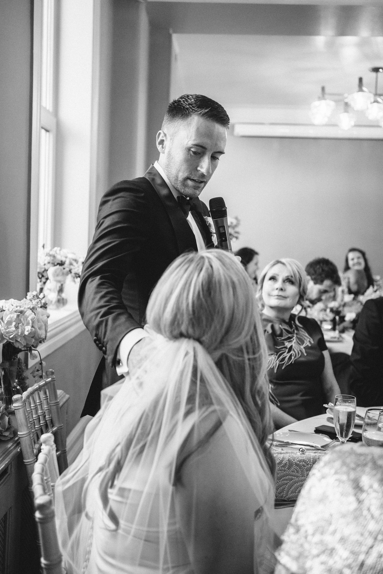 A speaker in a dark suit addresses wedding guests during an emotional reception moment.