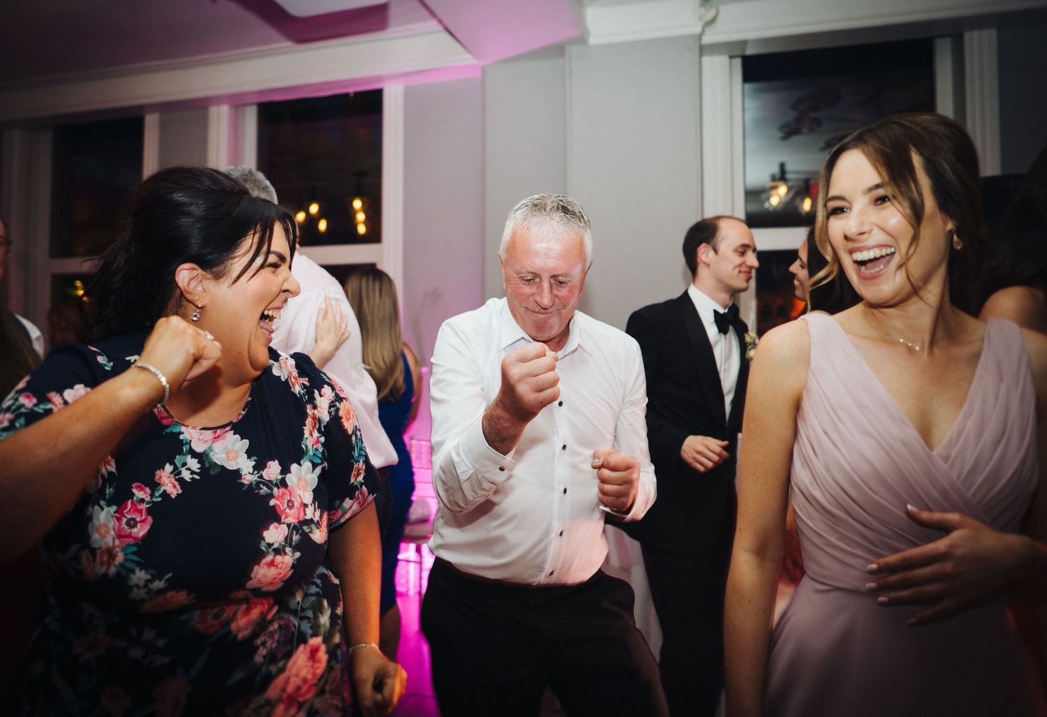 Party guests laughing and dancing together under pink lighting at a wedding reception.