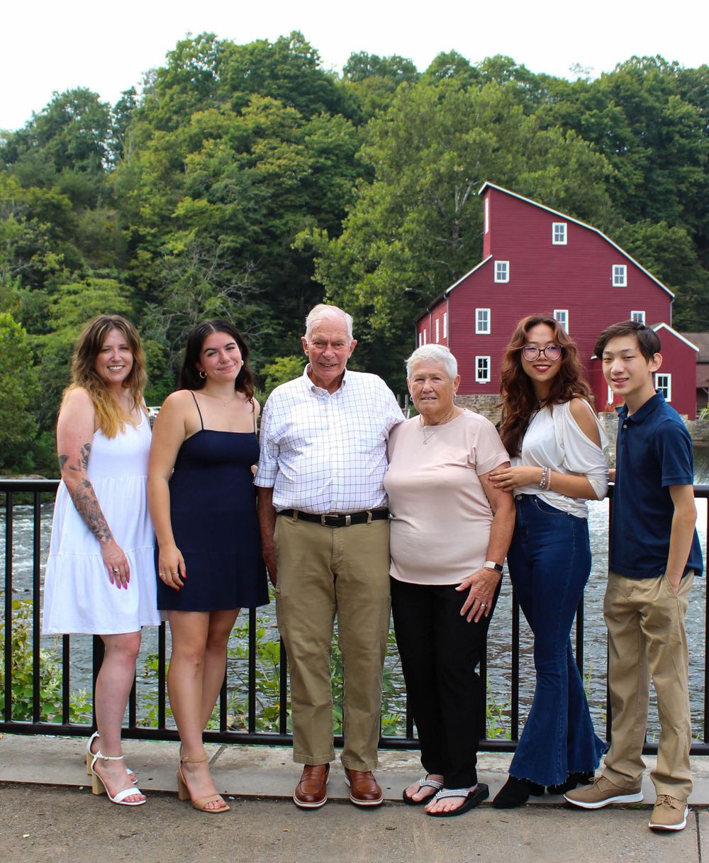 A multi-generational group poses on a walkway with a classic red mill building in the background.