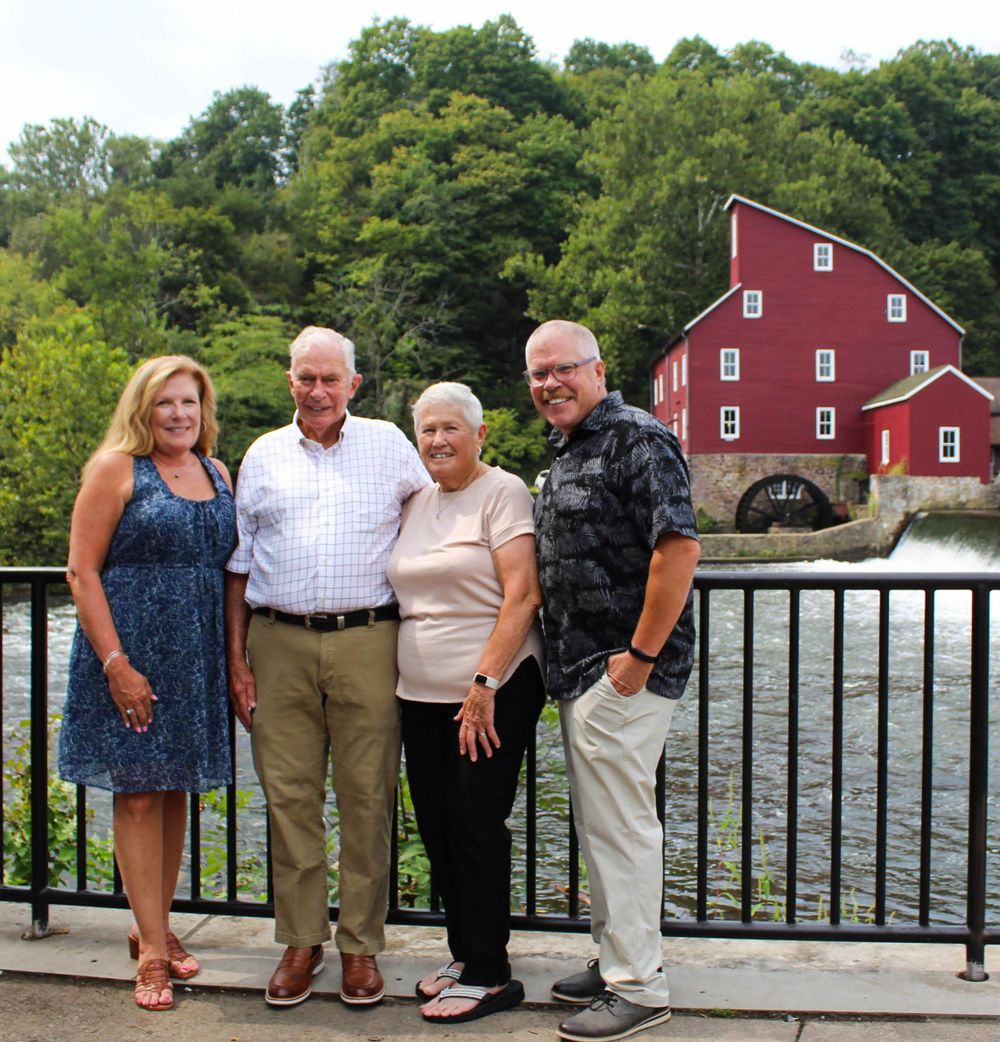 A group stands together on a deck overlooking a red mill building and pond on a sunny summer day.
