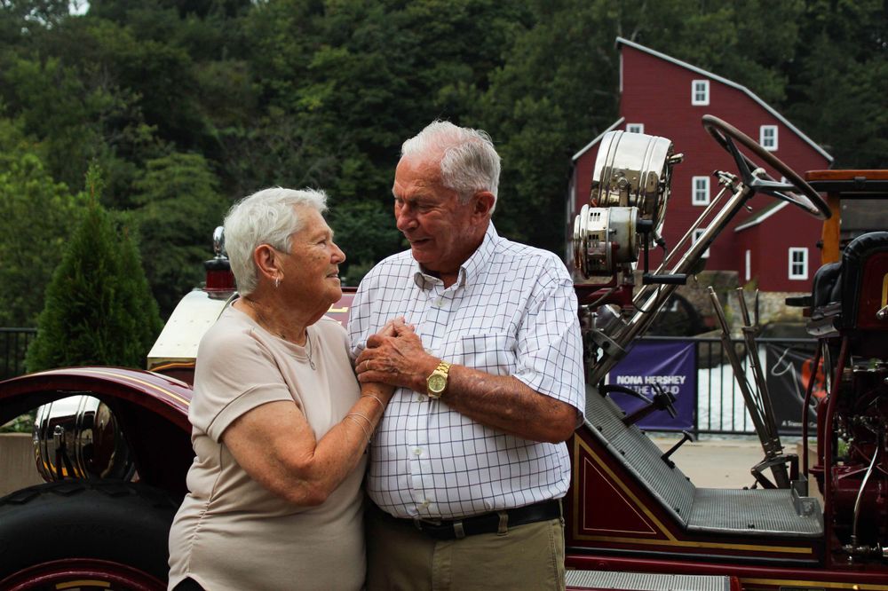An elderly couple shares a sweet dance moment in front of a red barn structure.