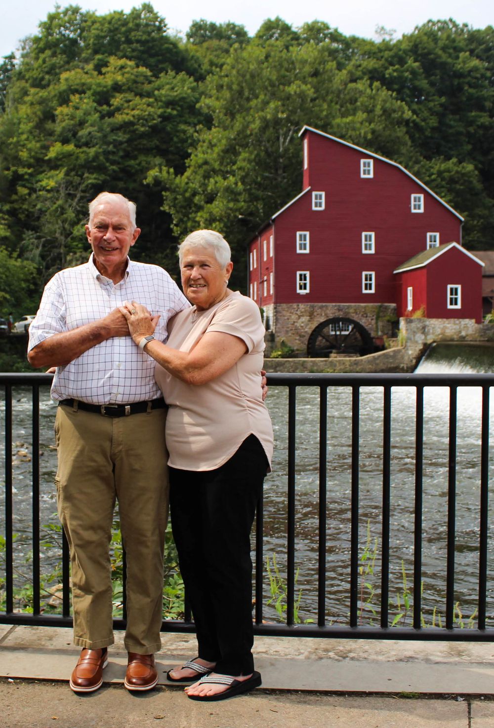 Two visitors pose at viewing area with Red Mill Museum and waterfall behind them.