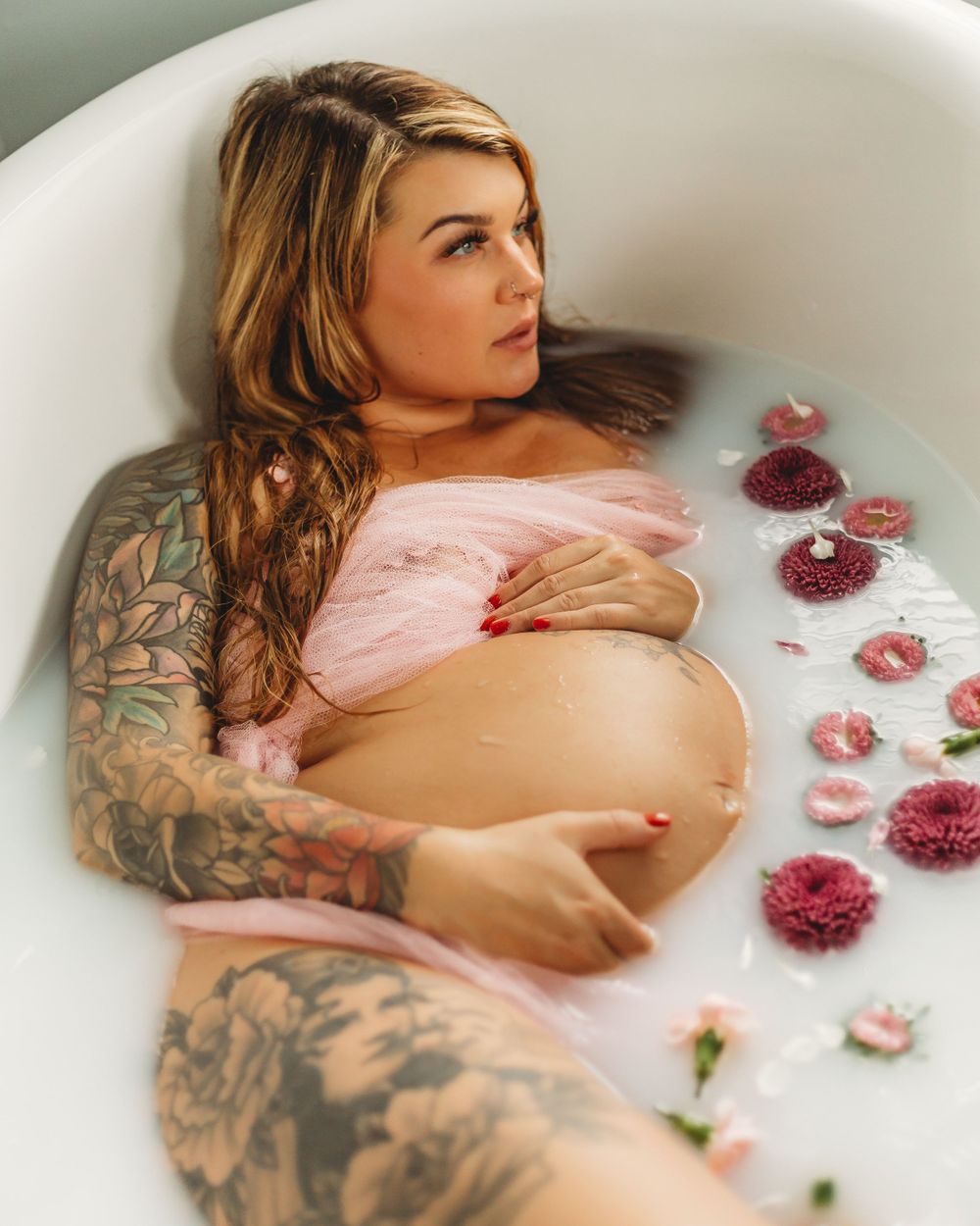 Dreamy maternity milk bath scene with pink fabric and deep red flowers floating on water.