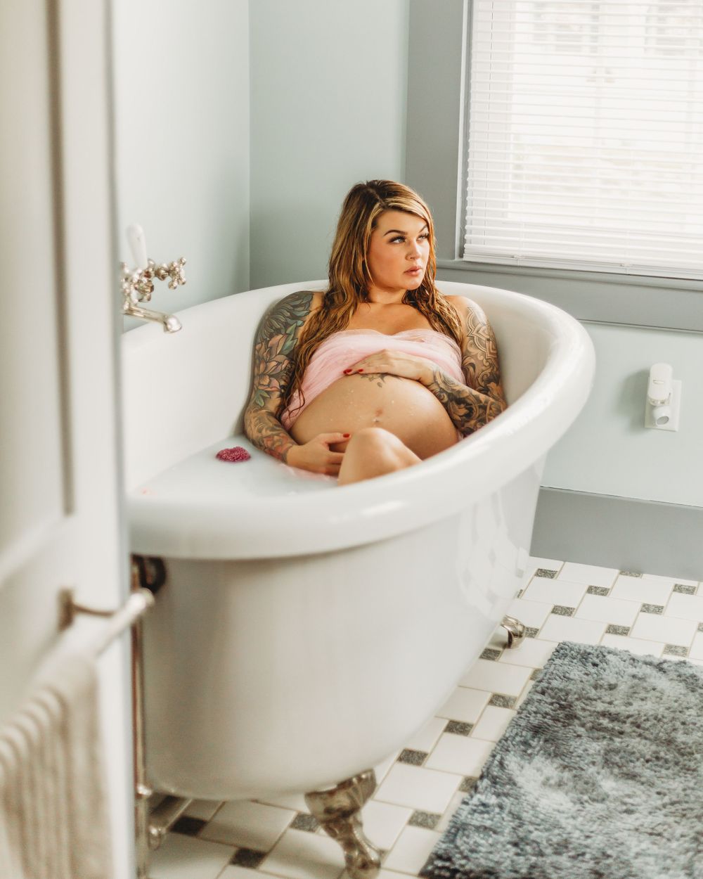 Vintage-style clawfoot tub maternity photo with checkered floor tiles and natural lighting.