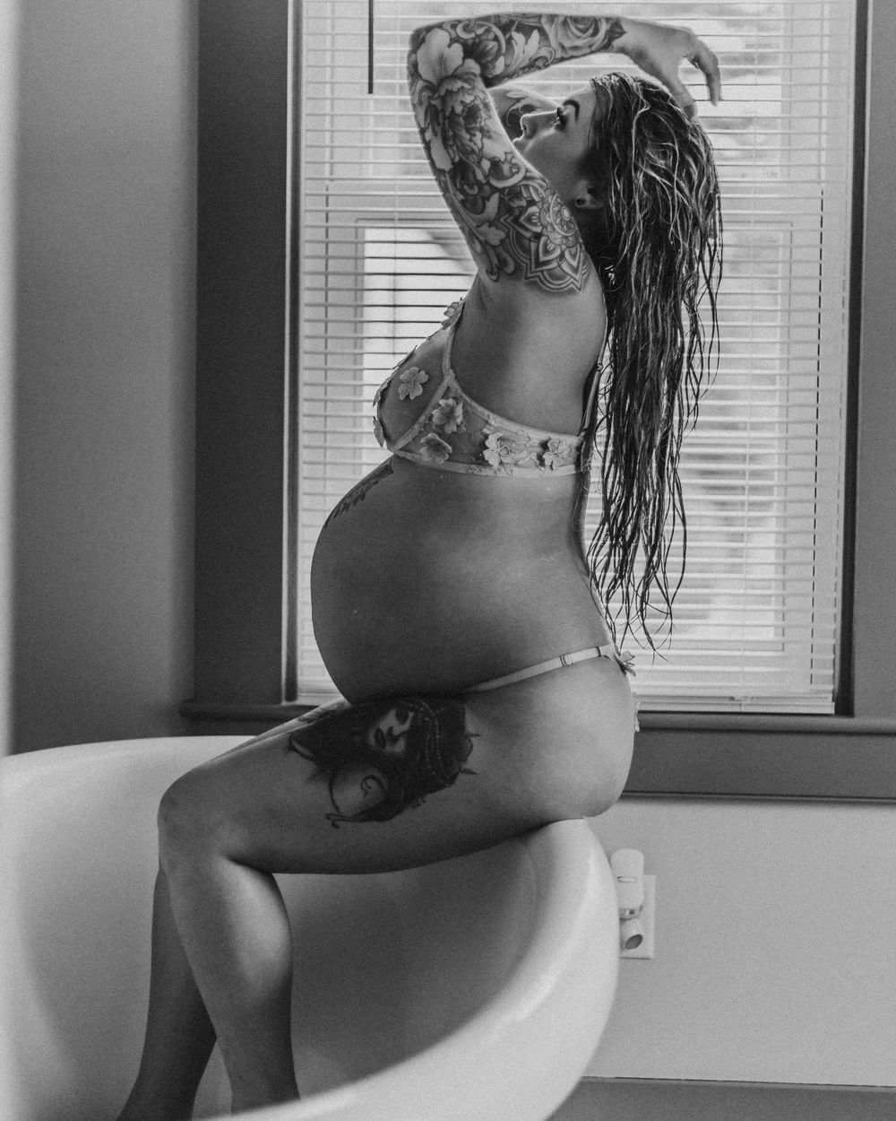 Black and white photo of a pregnant person with tattoos posing on a bathtub ledge near a window.
