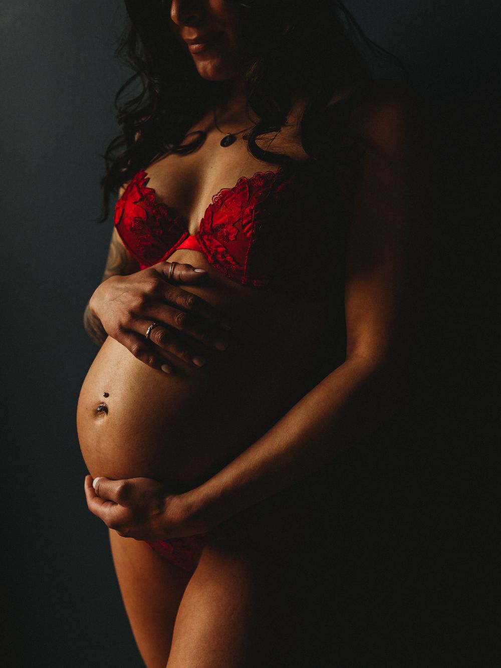 Intimate maternity portrait with red lingerie in moody lighting.
