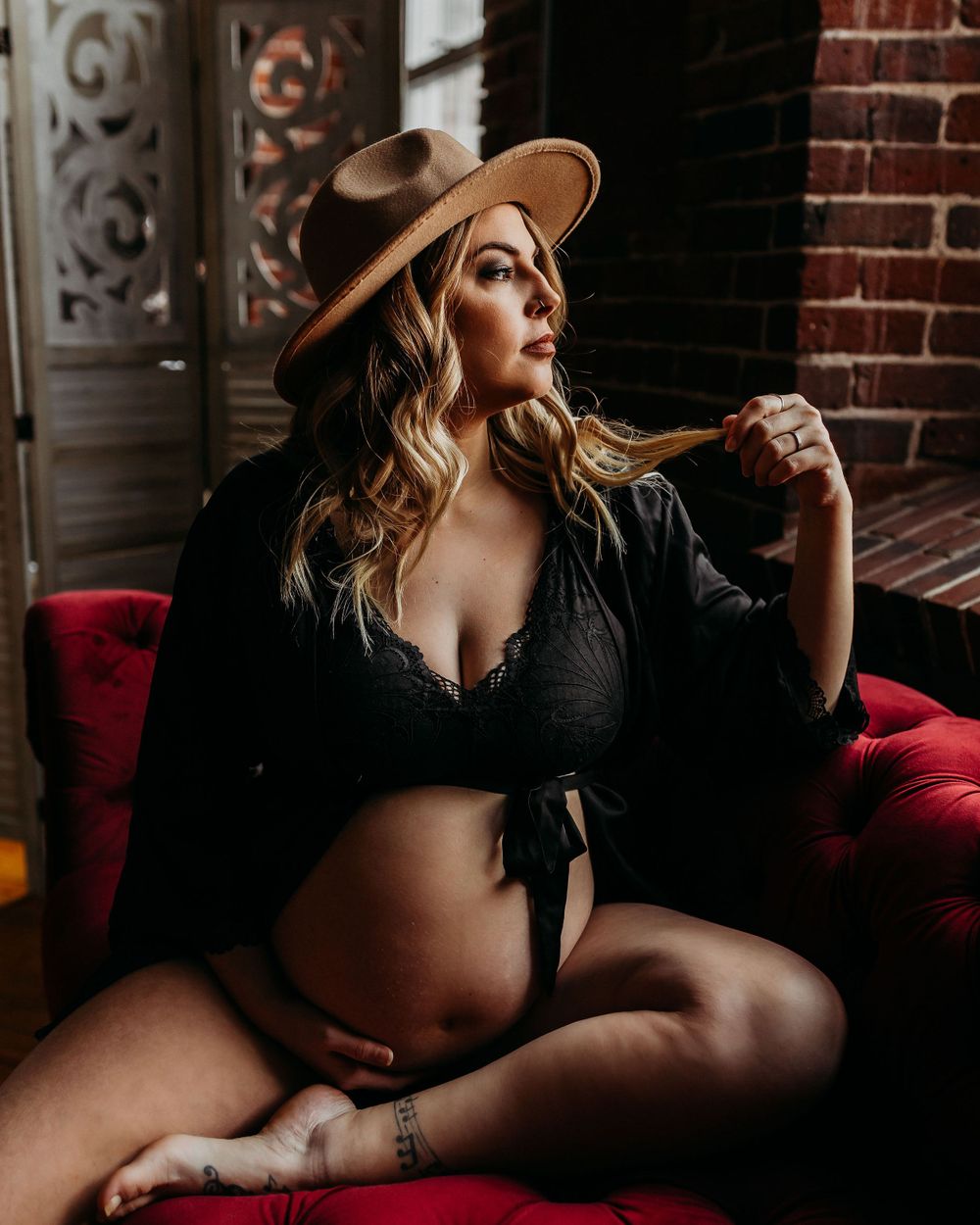 Moody maternity boudoir photo on red velvet couch with brown hat and black wrap.