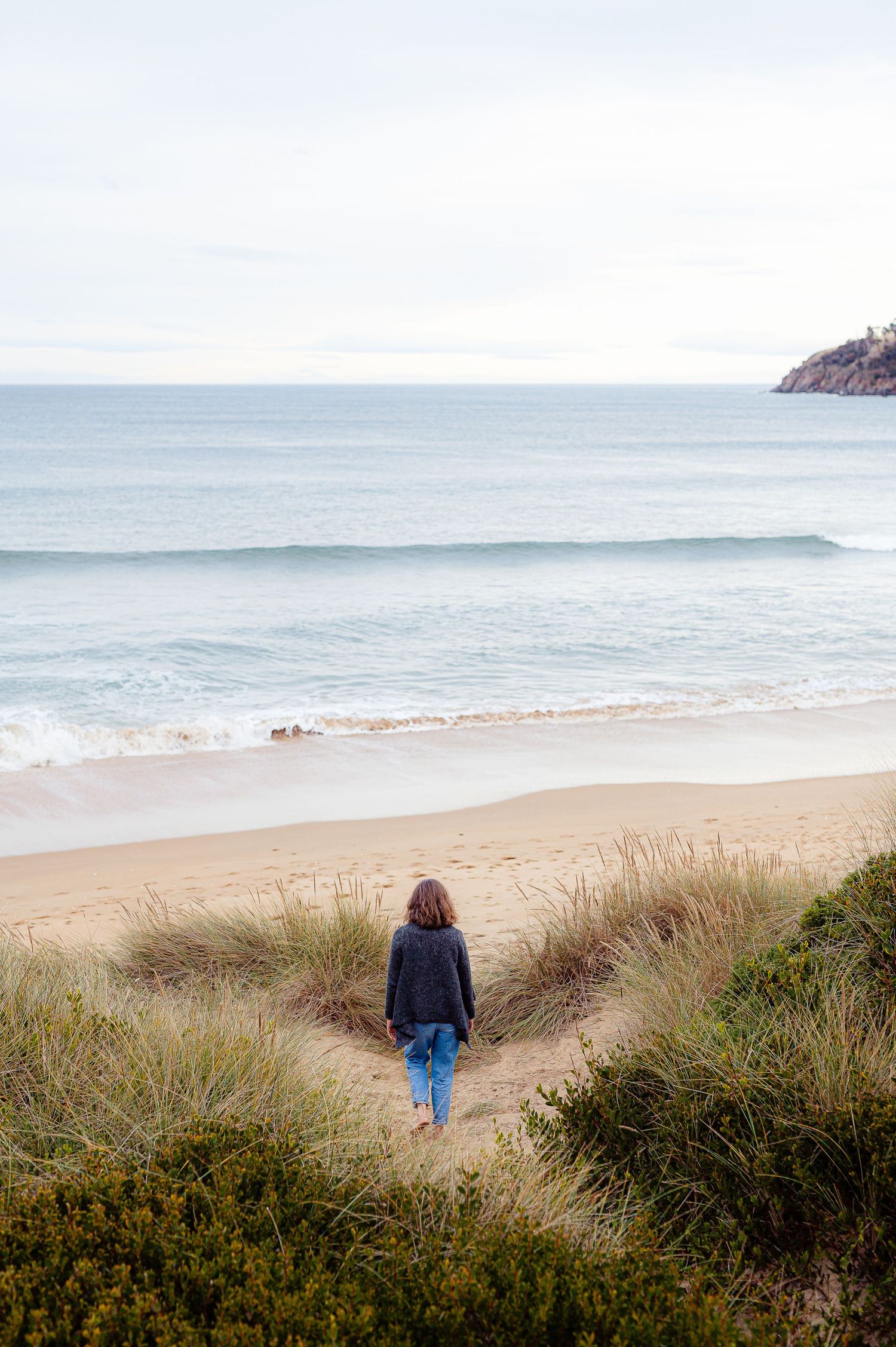 A solitary figure walks down sandy path toward a misty beach shoreline.