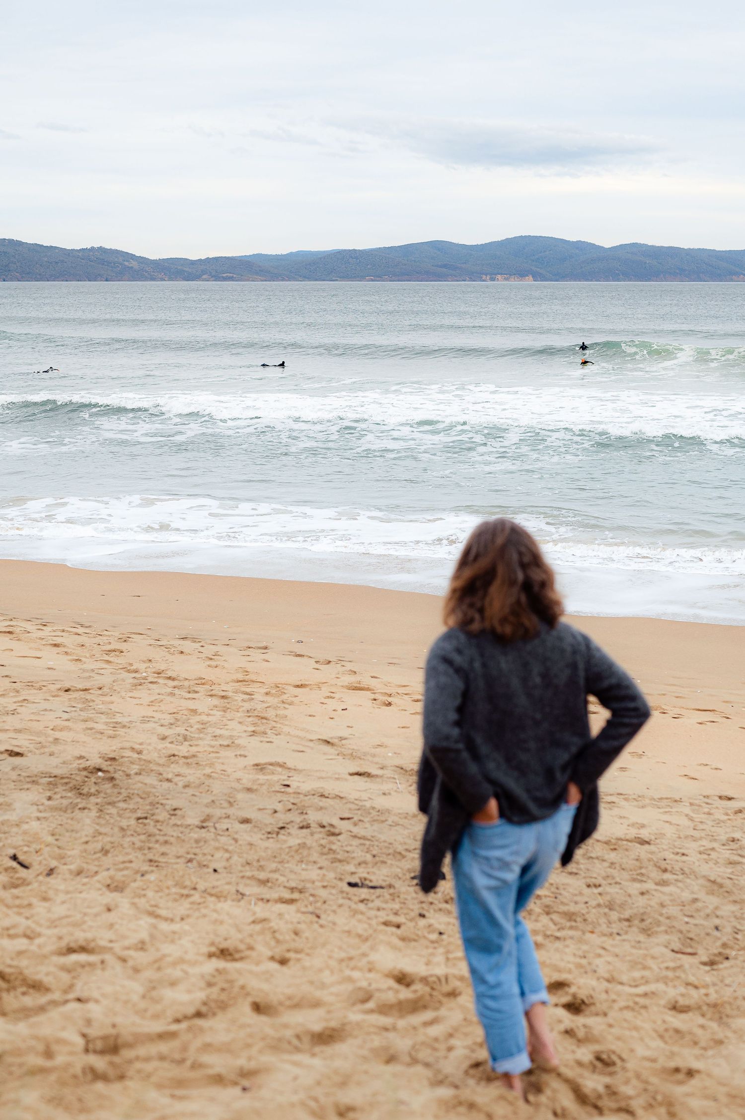 A person in a sweater and jeans stands on a beach watching surfers ride waves with mountains in the distance.