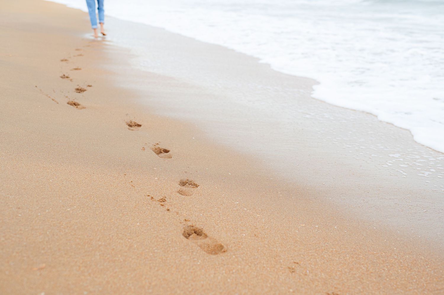 Footprints trail along the wet sand of a beach as waves wash up on shore.