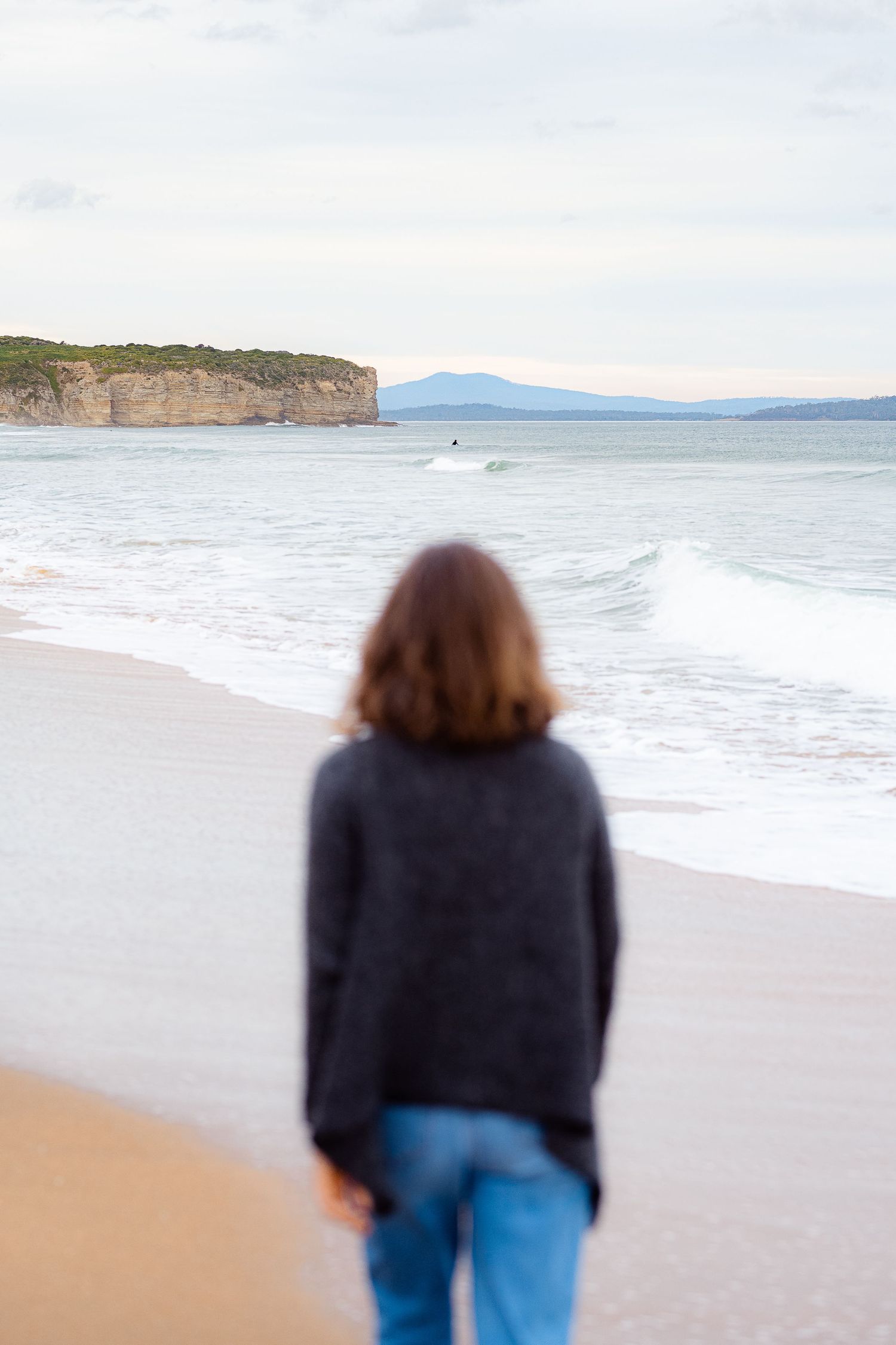 A person in blue jeans and dark sweater looks out at cliffs and ocean waves from sandy beach.