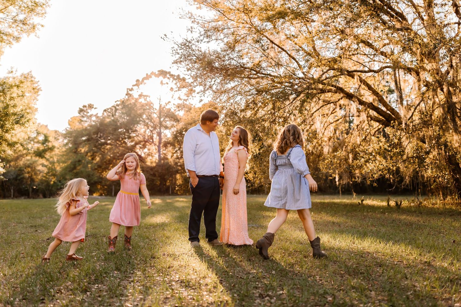 Family of five walking together in a sunlit park with large oak trees in the background.