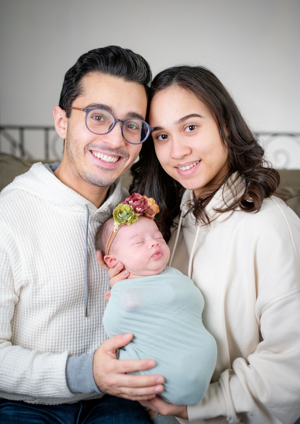 Young couple in neutral tones holding their swaddled newborn with floral headband.