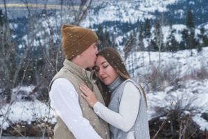 Couple embracing tenderly in snowy mountain landscape, man kissing woman's forehead during winter snowfall.