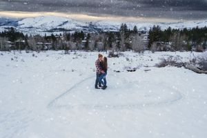 Couple embracing in snowy field with heart drawn in snow, surrounded by winter landscape.
