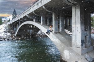 Person sitting alone under concrete bridge arch over river during winter snowfall.