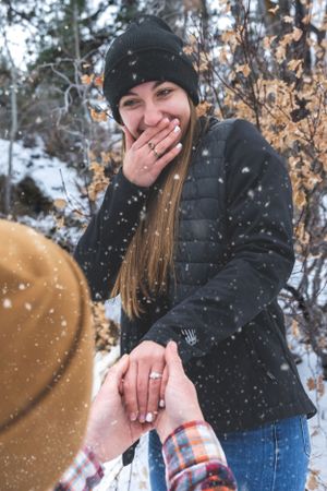 Woman laughing with hand over mouth during snowy winter marriage proposal, wearing black jacket and beanie hat.