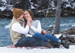 Happy couple sitting on snowy ground laughing together beside a winter river during snowfall.