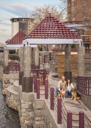 A couple with two dogs poses near a stone waterway with a pyramid-roofed gazebo in a riverside park.