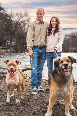 A happy couple walks two dogs along a rocky riverbank at sunset in an outdoor portrait.