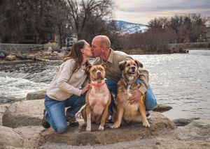A couple kisses while crouching between their two dogs on a rocky riverbank at dusk.