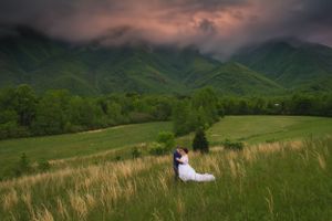 Bride and groom embracing in a lush green meadow with dramatic storm clouds over misty mountain backdrop.