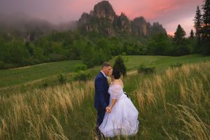 Wedding couple holding hands in tall grass field with foggy rocky mountains at pink sunset.