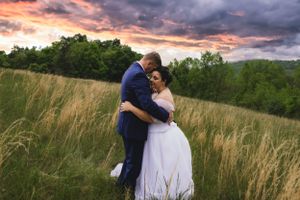 Bride and groom sharing a romantic embrace in golden grass field under a vibrant dramatic sunset sky.
